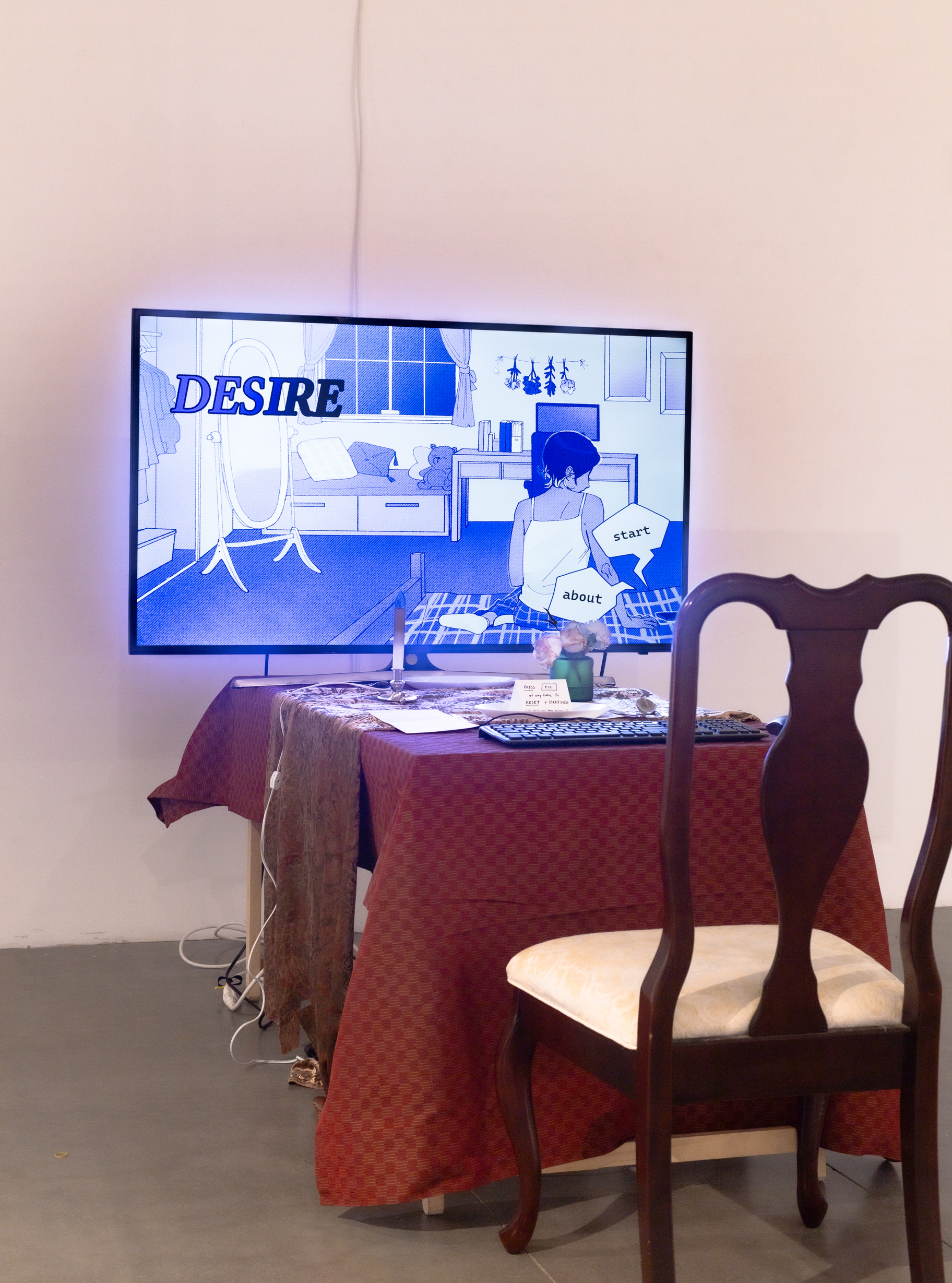 An upholstered chair sits in front of a table with tablecloth. On top of the table is a keyboard, a computer mouse, another table runner, a glass case with two flowers, a fake wax candle, and a television. On the TV is the start screen of an interactive game.